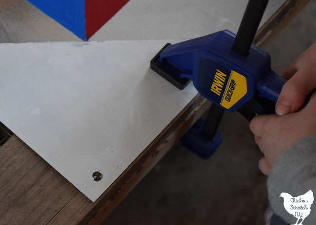 hole drilled in sheet metal barn quilt being held down by a quick grip clamp to a scrap of wood