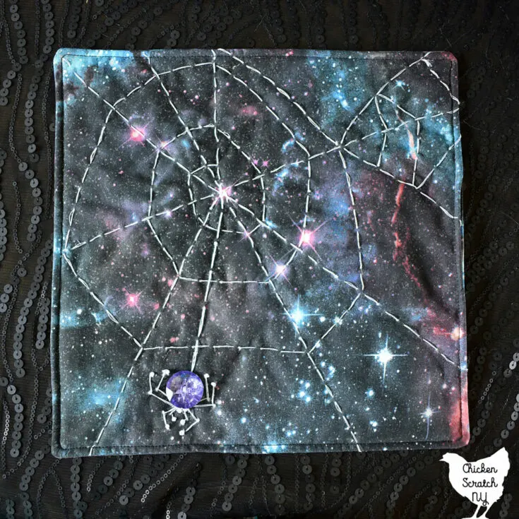 small square spiderweb mini quilt with hand embroidery on galaxy print fabric with a crystal button spider