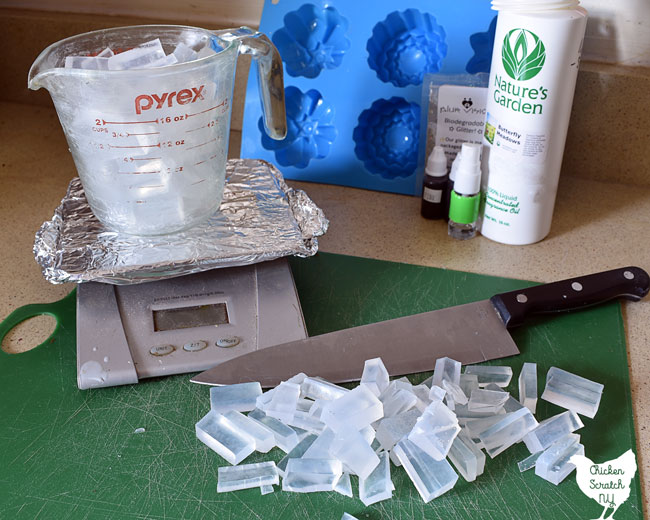 food scale covered in aluminum foil holding a glass measuring cup filled with chunks of clear melt and pour soap base on a green plastic cutting board with a knife and more pieces of chopped up soap base