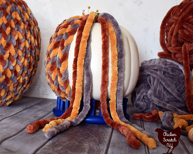 fake pumpkin held up with a blue stand being covered with braids made crom chunky chenille home yarn