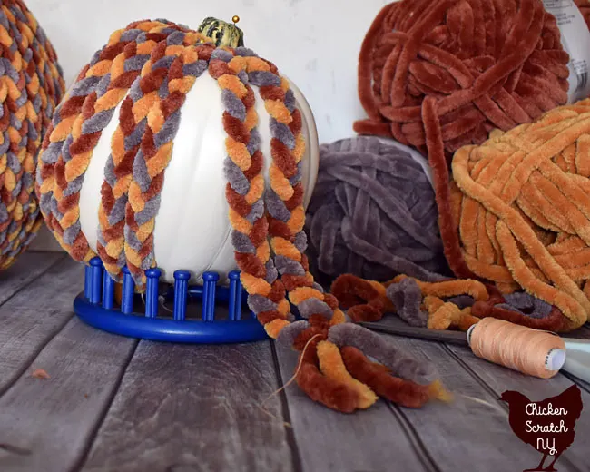 fake pumpkin held up with a blue stand being covered with braids made from chunky chenille home yarn being tied together to stop from unraveling