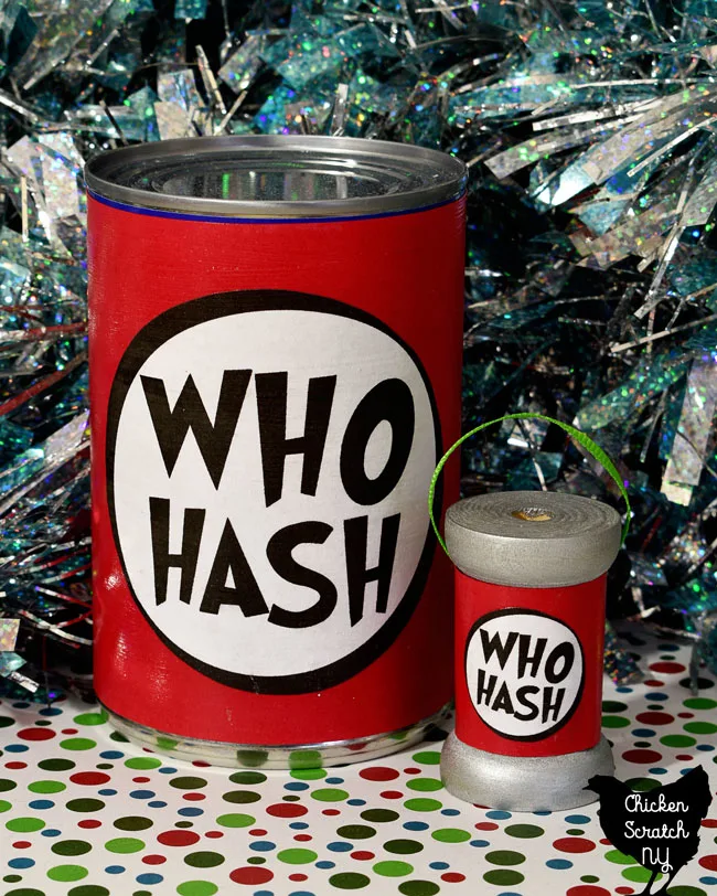 standard sized tin can with black, red and white "Who Hash" label next to a wooden spool with matching label and green ribbon hanging loop on a red, green and white paper with sparkly tinsel background