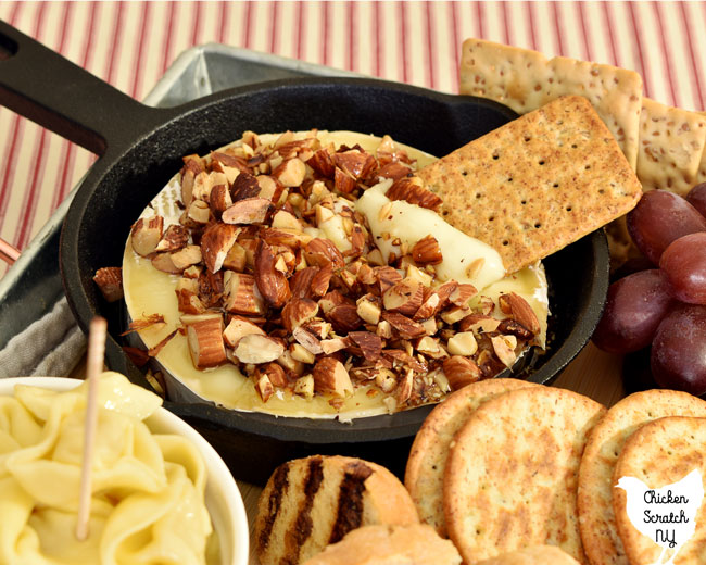 small cast iron skillet with baked brie topped with chopped almonds and maple syrup in a charcuterie tray