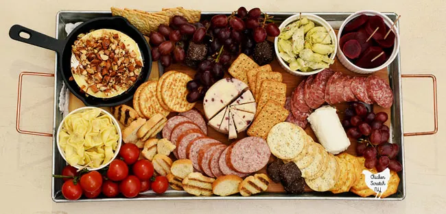 top view of low cost charcuterie board with baked brie, pickled beets, white cranberry cheddar, goat  cheese, summer sausage, salami, red grapes, cherry tomatoes, crackers and dark chocolate caramels