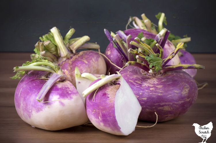 3 full purple top turnips on a wooden surface with one turnip sliced to show the white interior 