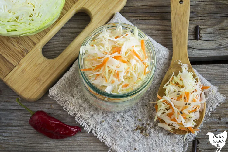 glass mason jar filled with shredded cabbage and carrots on a wooden cutting board with a hot hepper and half a cabbage