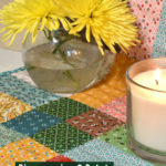 disappearing nine patch table runner made with Lori Holt Stitch fabric with a vase holding two yellow spider mums and a lit candle with text overlay "Disappearing 9 patch table runner free quilt pattern"