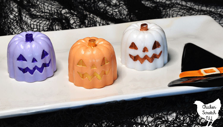 three soap pumpkins, one purple with dark purple face, one white with reddish copper face and one orange with gold face on a white plate with a witch hat decoration