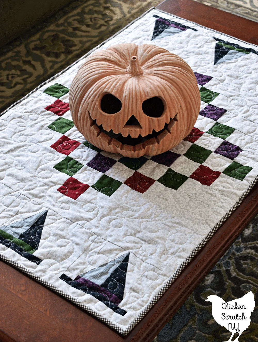 Halloween table runner made with red, green and purple accent fabric and witch hat quilt blocks with a large terra cotta pumpkin