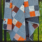 alfred quilt made with Halloween fabric hanging on a metal fence with text overlay stating "Alfred Quilt Pattern Free magic 4 patch PDF"