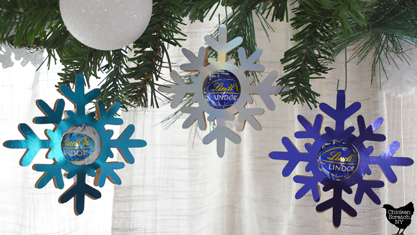 three cardstock snowflakes cut from metallic foil paper, each holding a blue wrapped Lindt truffle