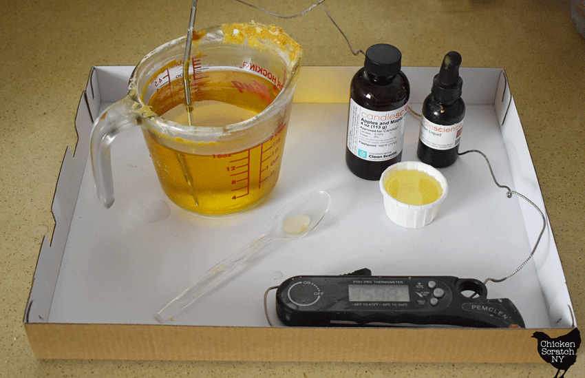 melted wax in a glass measuring cup with a thermometer sitting in a cardboard box lid to protect the table with a small whirte cup holding a yellow frangrance oil, a larger black bottle of fragrance oil and a small black bottle of candle dye with a dropper lid